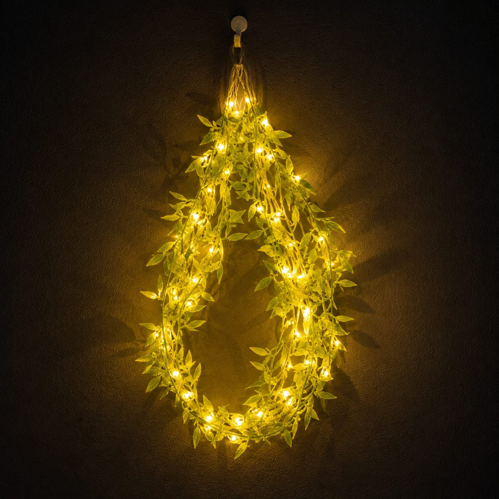 Warm white LED fairy string lights wrapped in artificial green leaves, arranged in a teardrop shape against a dark textured wall.