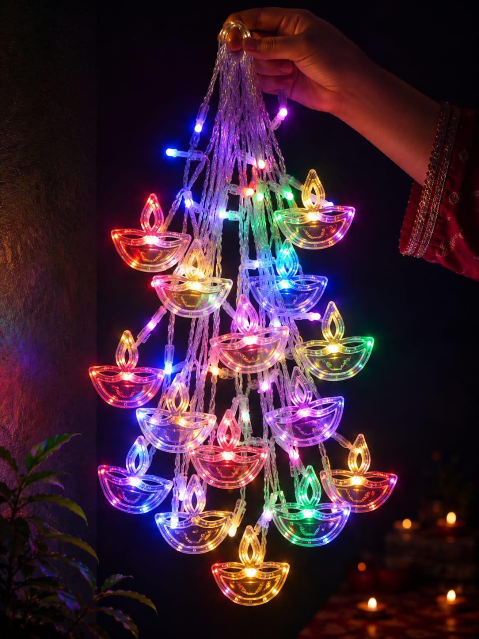 A hand holds a cluster of multicolor LED diya string lights shaped like oil lamps, glowing brightly against a dark background with warm bokeh lights in the distance.