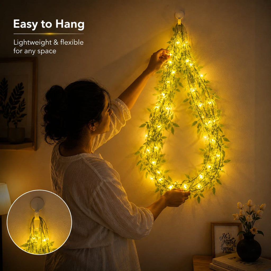 A woman hangs a flexible vine-style string light decoration shaped like a teardrop on a wall. The decoration is covered in glowing warm yellow lights and green leafy vines.