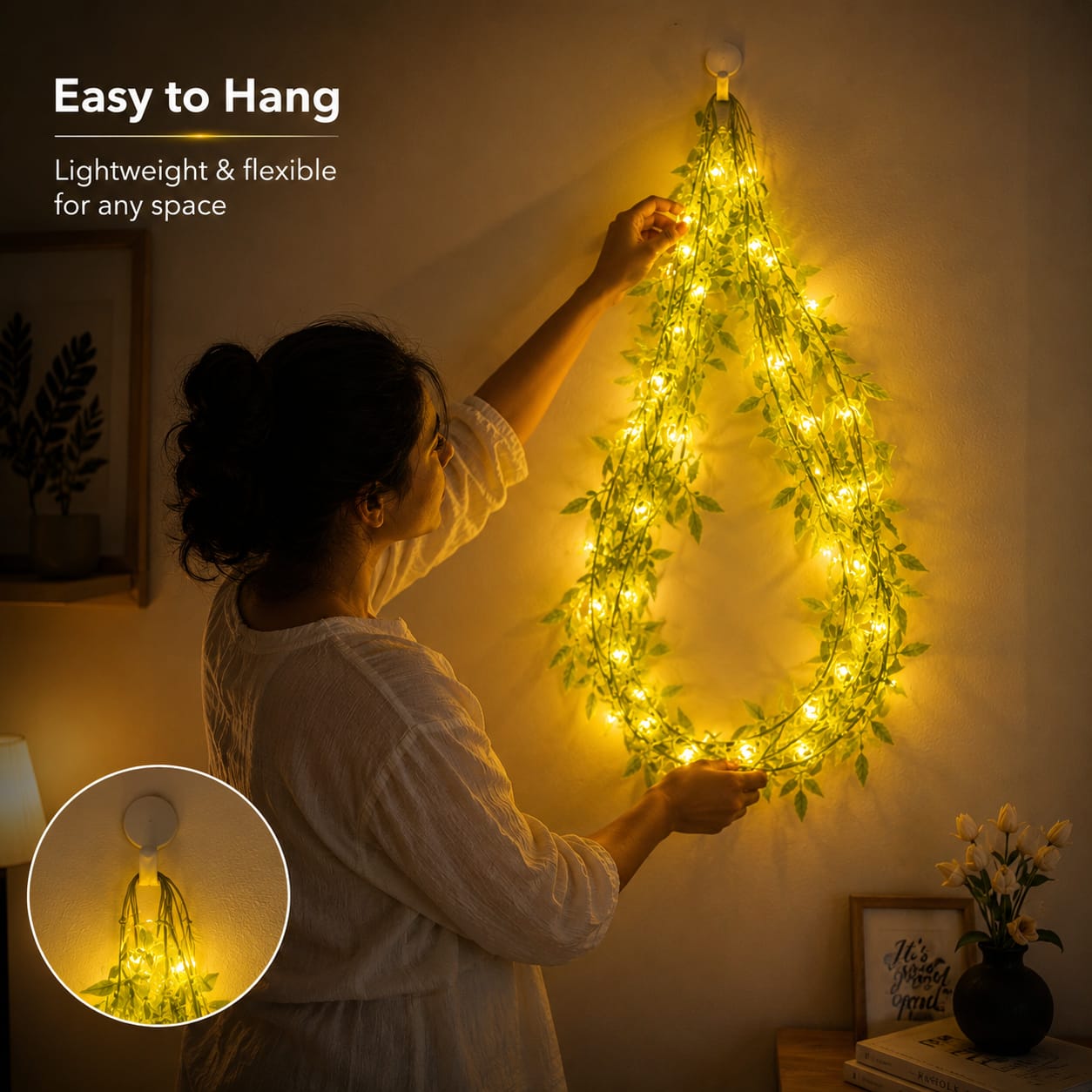 A woman hangs a flexible vine-style string light decoration shaped like a teardrop on a wall. The decoration is covered in glowing warm yellow lights and green leafy vines.
