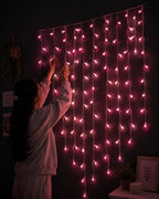 A person in a light-colored sweatshirt adjusts pink curtain string lights hanging on a dark wall in a cozy room setting.