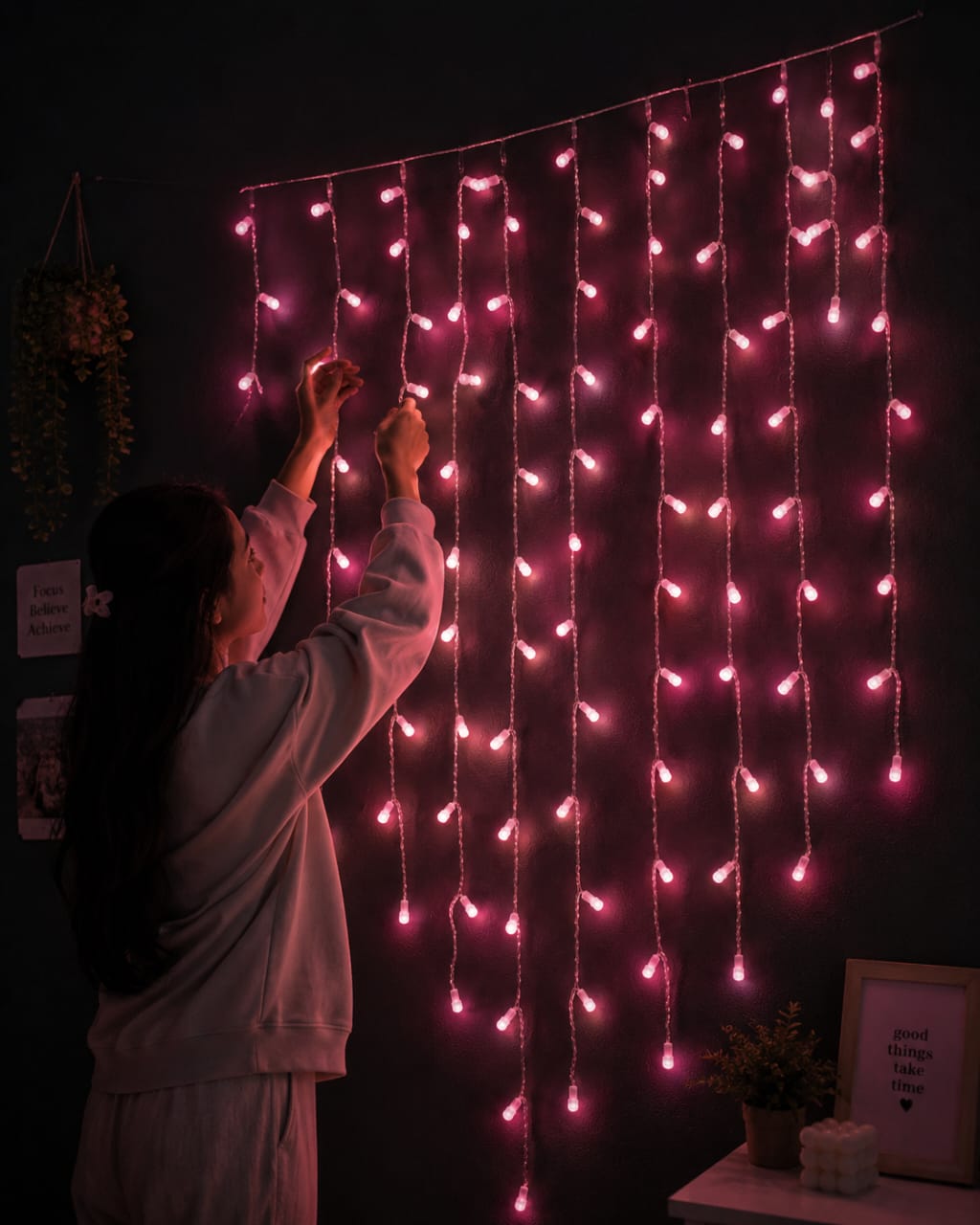 A person in a light-colored sweatshirt adjusts pink curtain string lights hanging on a dark wall in a cozy room setting.