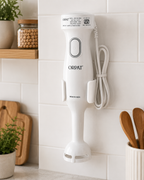 A white ORPAT immersion blender with a cord, mounted on a white tiled kitchen wall next to wooden cutting boards and a plant.